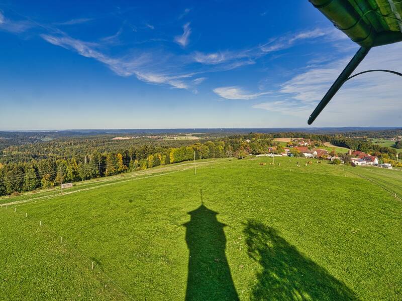 Eine weite grüne Wiese erstreckt sich vor einem Panorama aus Wäldern und einem Dorf unter einem klaren blauen Himmel.