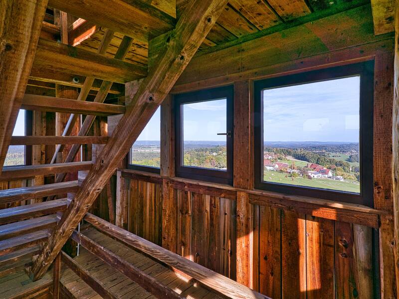Holztreppen und Fenster in einem Aussichtsturm bieten einen Ausblick auf eine ländliche Landschaft mit Feldern und Gebäuden.