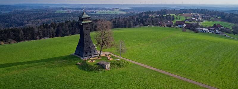 Ein schwarzer Aussichtsturm steht auf einem weiten, grünen Feld, umgeben von Bäumen und in der Ferne sind kleine Gebäude sowie ein Wald zu sehen.