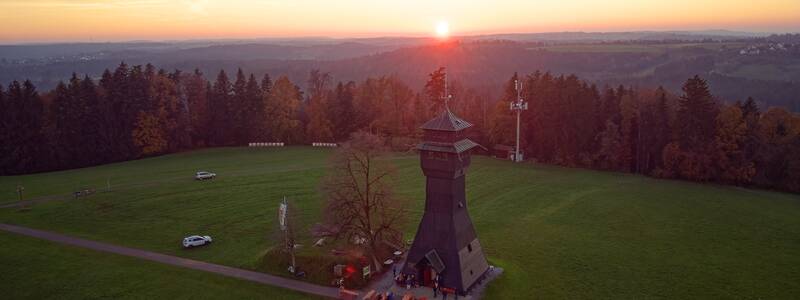 Ein einzelner Holzturm steht auf einer weitläufigen grünen Wiese, umgeben von Wald, während am Horizont die Sonne untergeht.