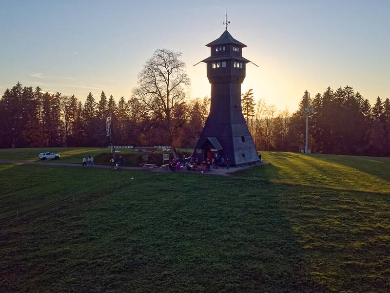 Eine Gruppe von Menschen versammelt sich bei Sonnenuntergang um einen Holzturm in einer Wiese, umgeben von Bäumen.