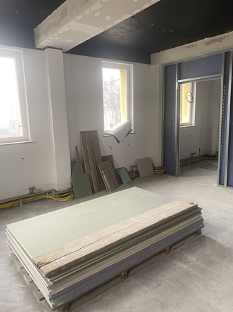 Ein unfertiger Raum mit zwei Fenstern, Gipskartonplatten auf dem Boden und Bauwerkzeugen an den Wänden.