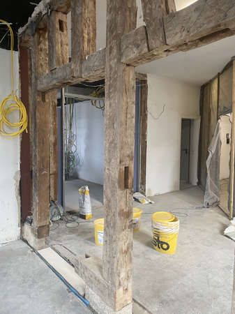 In einem Raum im Renovierungsprozess sind unverputzte Holzbalken, gelbe Eimer und Kabel an den Wänden sichtbar.