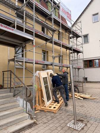 Zwei Männer arbeiten an einem Gebäude mit Gerüst, wobei sie Fensterrahmen auf Holzpaletten untersuchen oder bearbeiten.